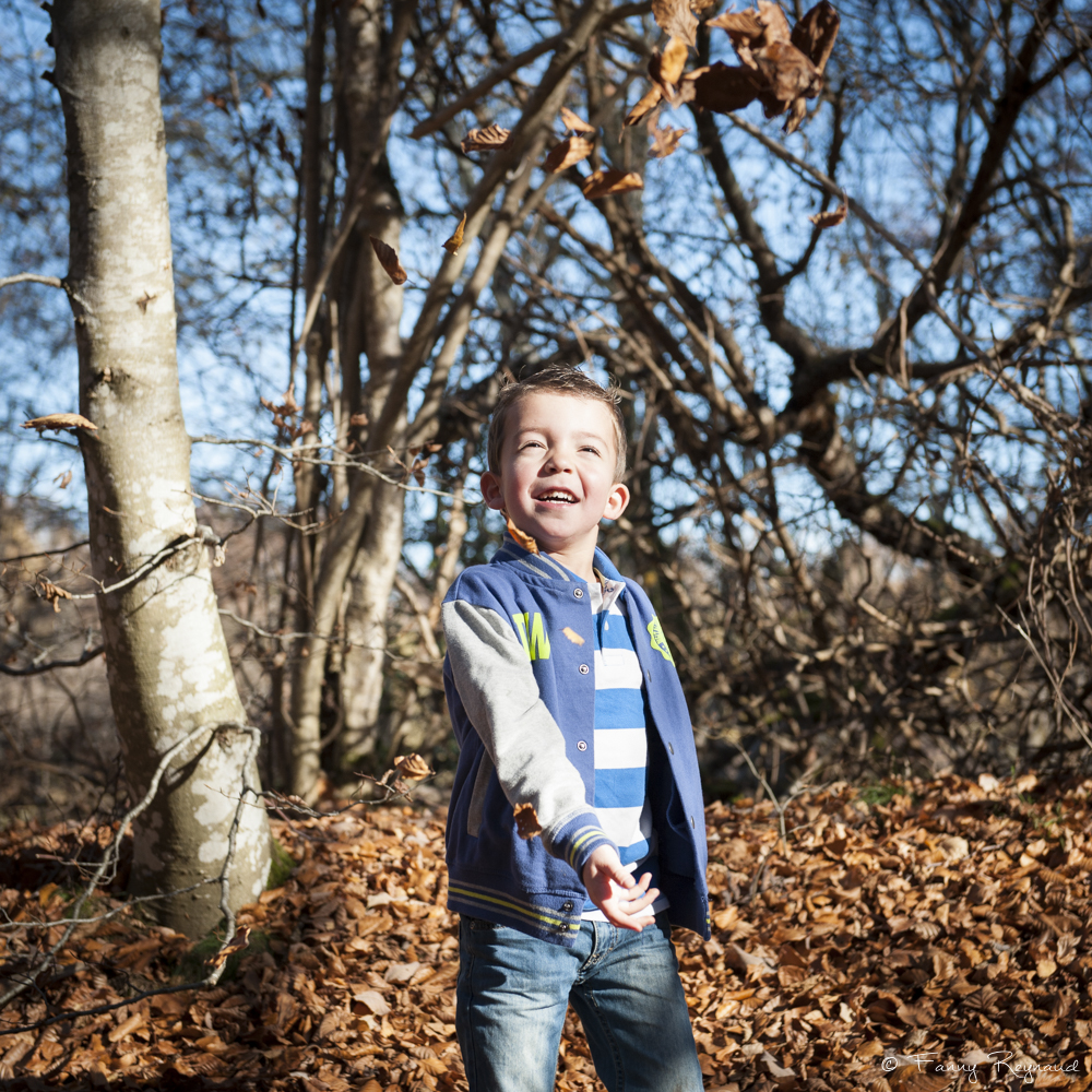 Portrait d'un petit garçon dans la forêt en train de jouer avec des feuilles d'automne. Image extraite d'un shooting en extérieur dans les volcans d'auvergne par fanny reynaud photographe professionnelle à clermont-ferrand.