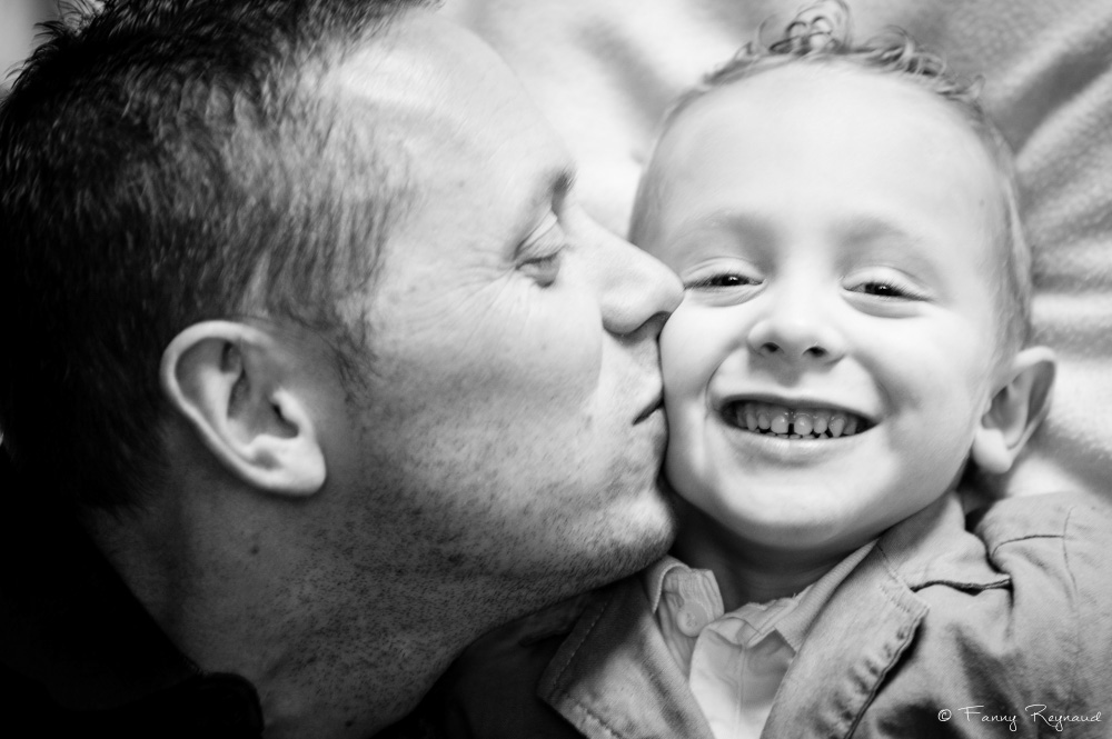 Photographie noir et blanc d'un papa embrassant son fils sur la joue lors d'une séance à domicile. © Fanny Reynaud photographe à domicile.