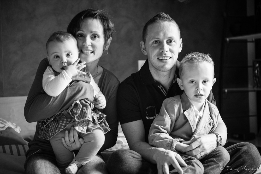 Photo de famille à domicile en lumière naturelle en noir et blanc : un jeune couple avec leurs deux enfants posent dans leur chambre familial. © Fanny Reynaud photographe à domicile autour de clermont-ferrand.