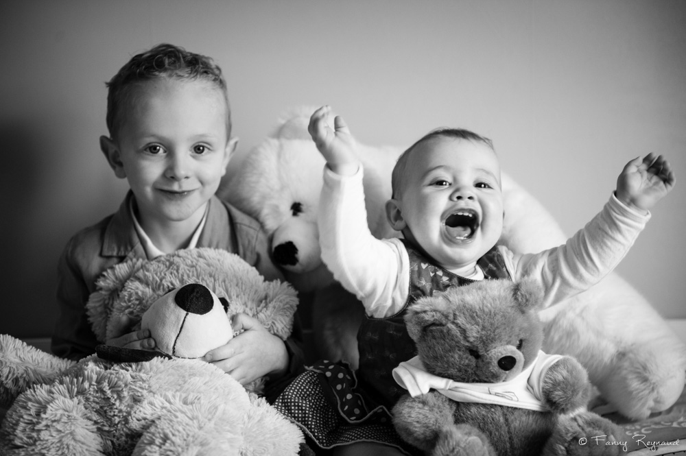 Photo de deux jeunes enfants souriant et jouant avec des peluches dans leur lit, extrait d'une séance à domicile dans les environs de riom, par une photographe professionnelle.