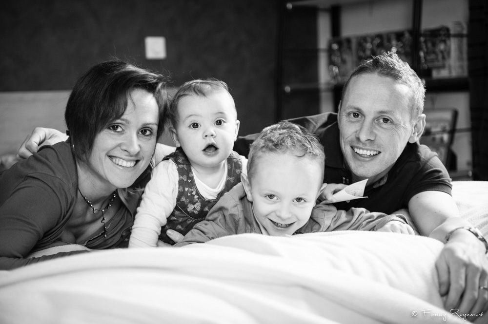 Photo de famille originale et décalée dans le lit familial : les parents et leurs deux enfants sourient. Extrait d'une séance de portrait à domicile par une photographe professionnelle de clermont-ferrand.
